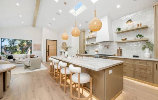Modern kitchen interior with wooden cabinets, island, and pendant lights, featuring open shelves and a cozy living area.